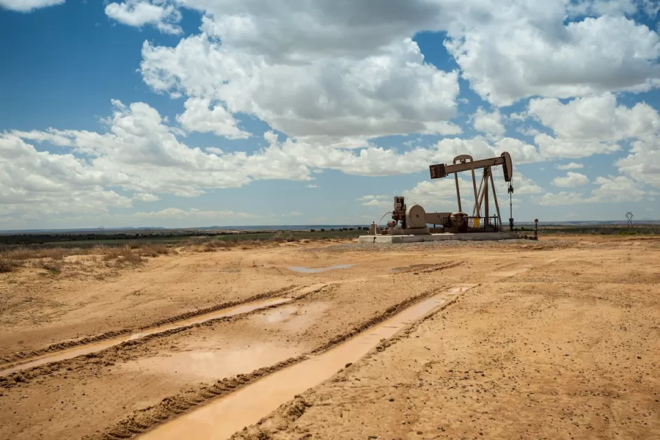 Distant view of brown oil pump jack in a flat desert with cloudy blue sky.